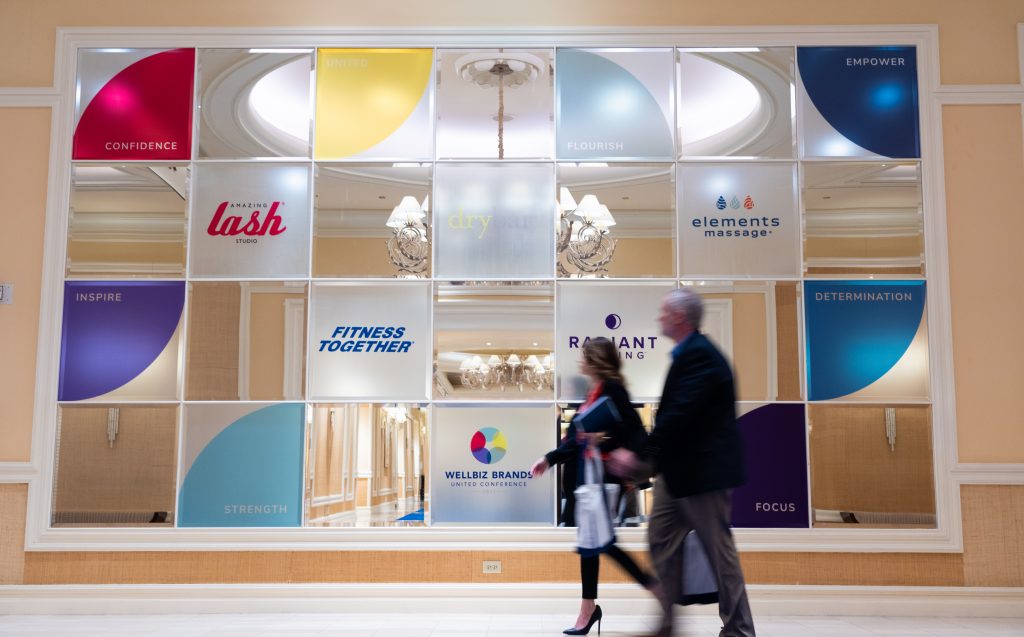 Attendees walking past wall displaying various WellBiz Brands