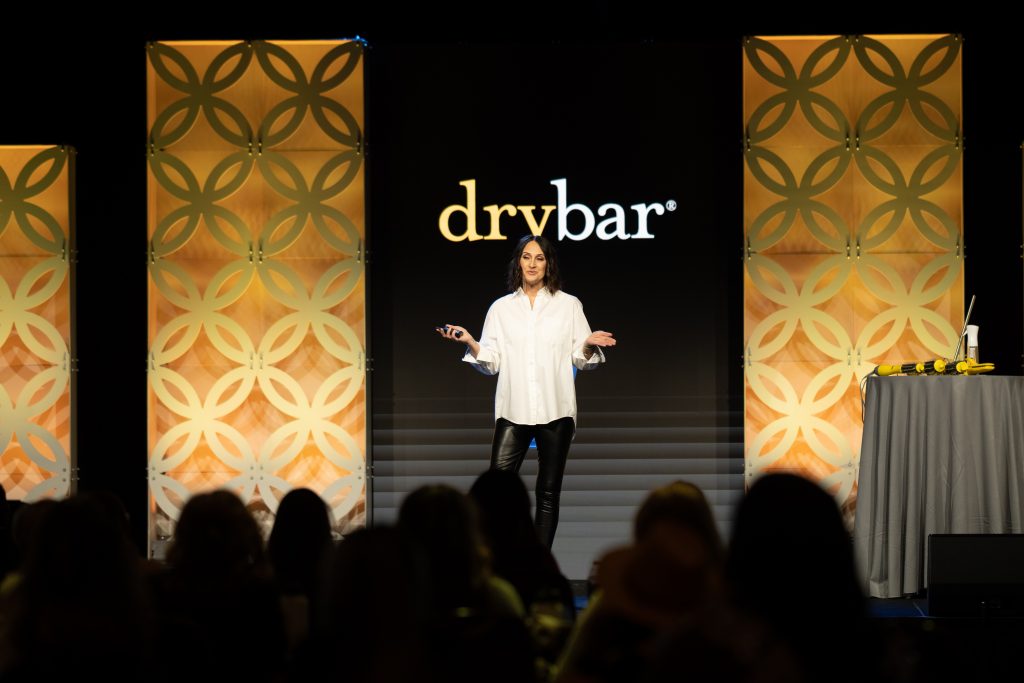 Speaker on stage in front of "Drybar" sign